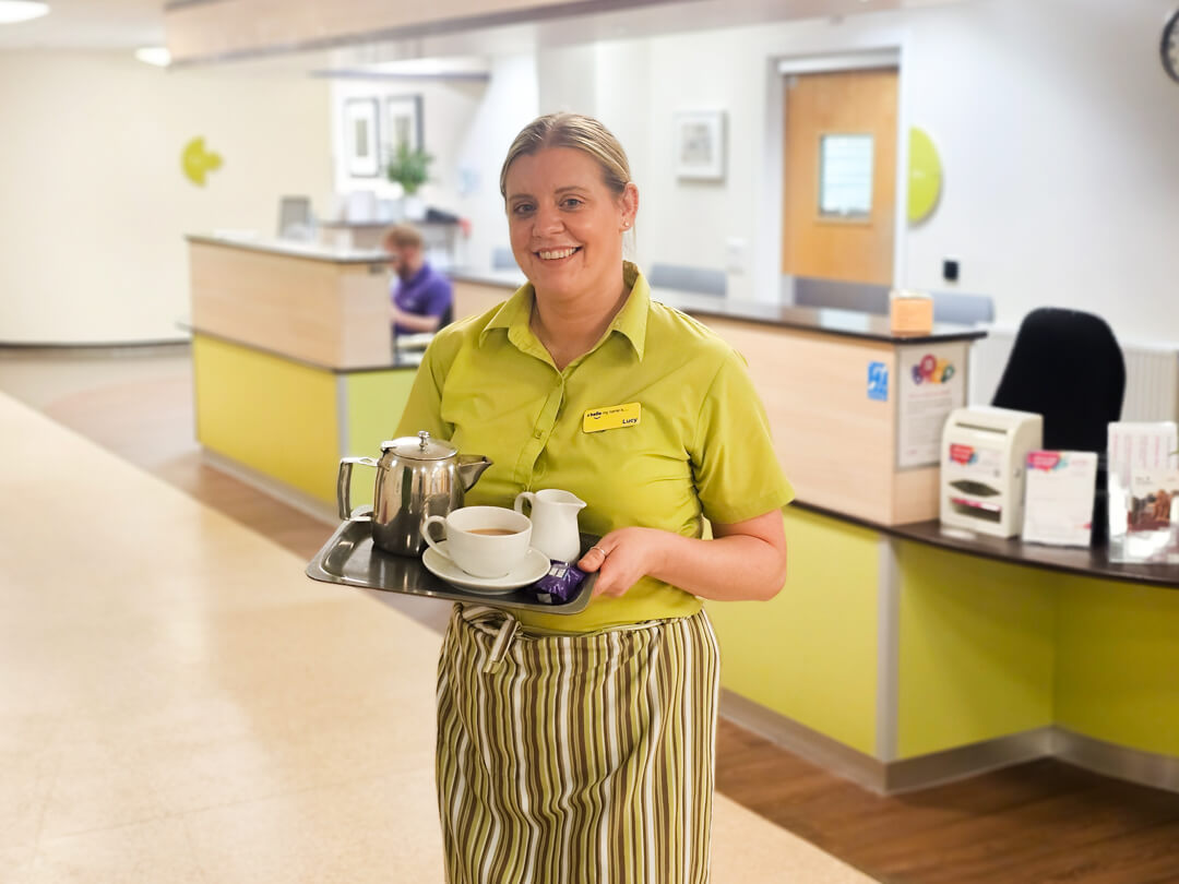 Lucy, a Hospitality Assistant at our In-Patient Centre, holding a tray with tea and biscuits, ready to serve patients and families.