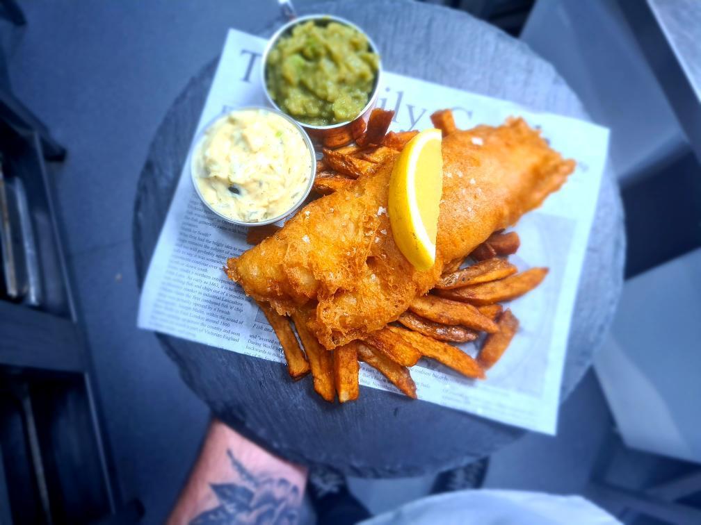 A photograph of fish and chips. A lemon wedge sits on top. It is accompanied by mushy peas and tartar sauce.