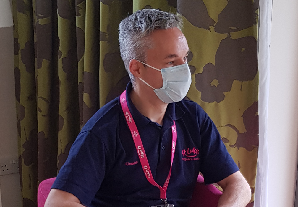 A photograph of our Chaplain Mark Newitt. He wears a face mask as he speaks to a patient, listening intently.