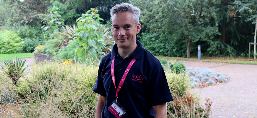 A portrait photograph of our Chaplain Mark Newitt. He smiles to the camera and stands in the St Luke's gardens. He is wearing a navy St Luke's branded polo shirt.